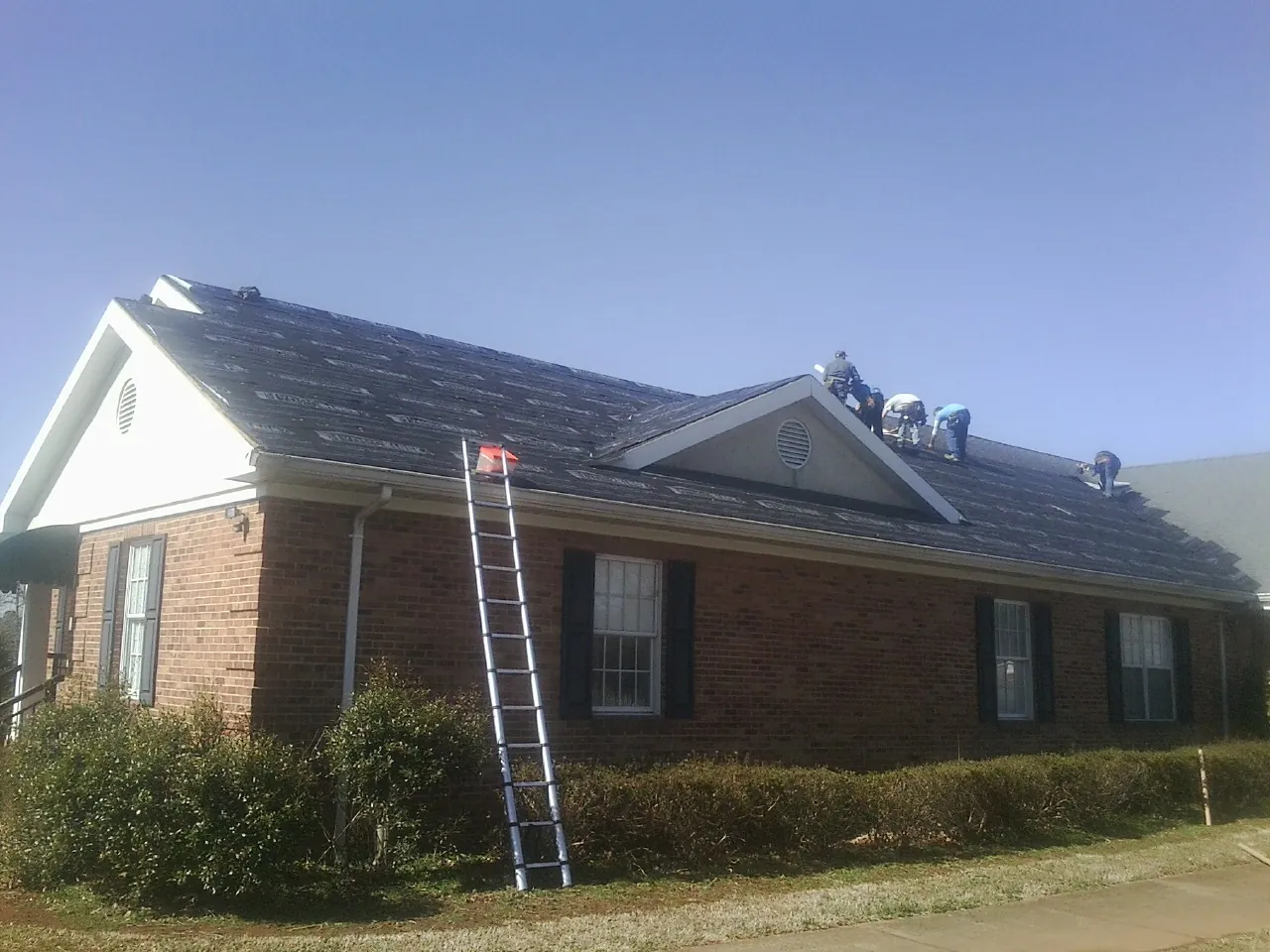 Crew tearing shingles off a brick ranch roof with a ladder set — Metal Roof Repair work by our crew in Glidden